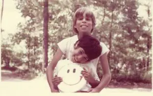 A young girl holding a smiley face pillow while another child looks on.