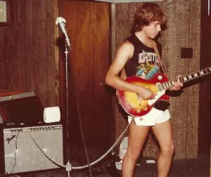 A man in shorts playing guitar and standing next to a microphone.